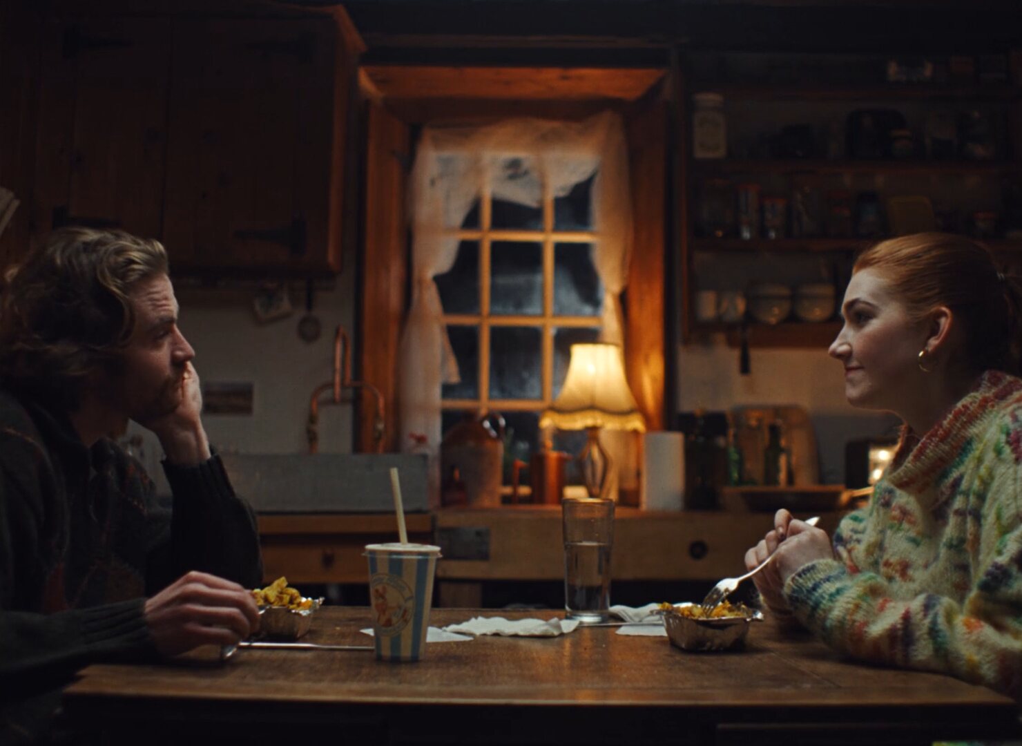 Two people sit across a kitchen table in a small Irish cottage, lit softly from above, with a condensation-covered window and a lit lampshade behind them.